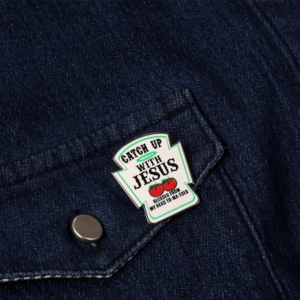 A Kiss Me Later enamel pin, shaped like a ketchup bottle on dark denim, reads Catch Up With Jesus in a classic label style, with tomato graphics and the phrase Blessed from my head to-ma-toes below.