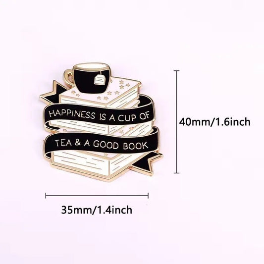 The Kiss Me Later enamel pin features a stack of books with a tea cup, wrapped in a banner reading Happiness is a cup of tea & a good book. Measures 40mm tall by 35mm wide.