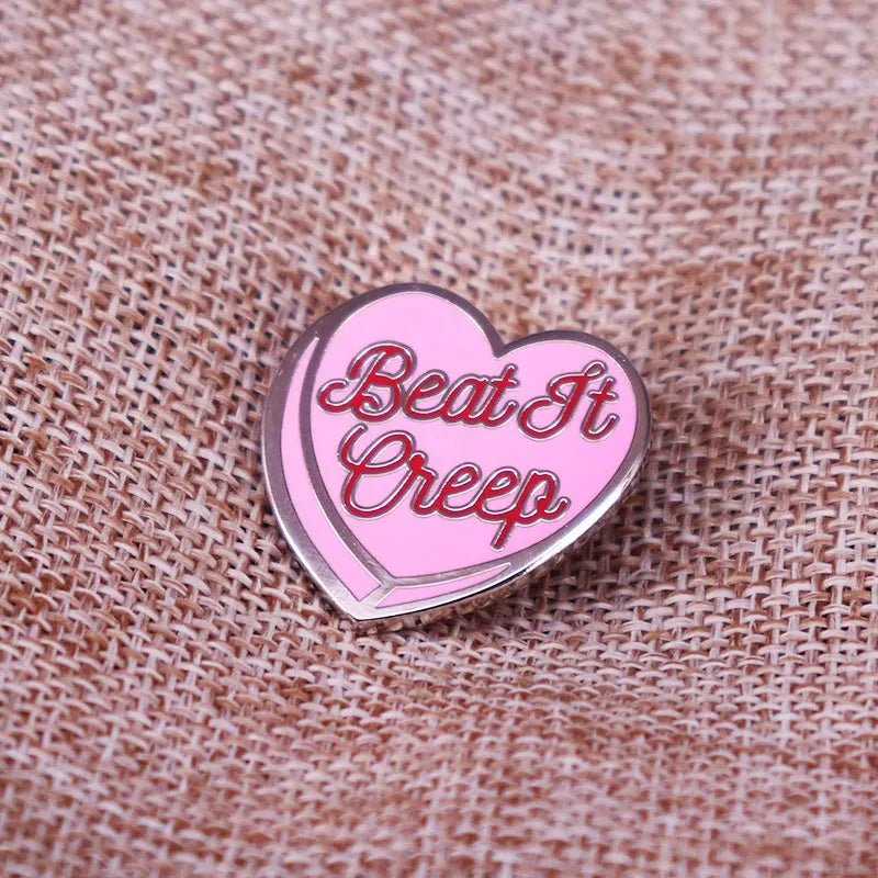 Pink and red enamel heart pin featuring Beat It Creep in red script, displayed against textured brown burlap fabric.