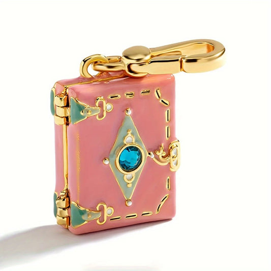 Small pink suitcase charm features gold accents, green corner details, a central blue gemstone reminiscent of an opening book, and is attached to a gold clasp.