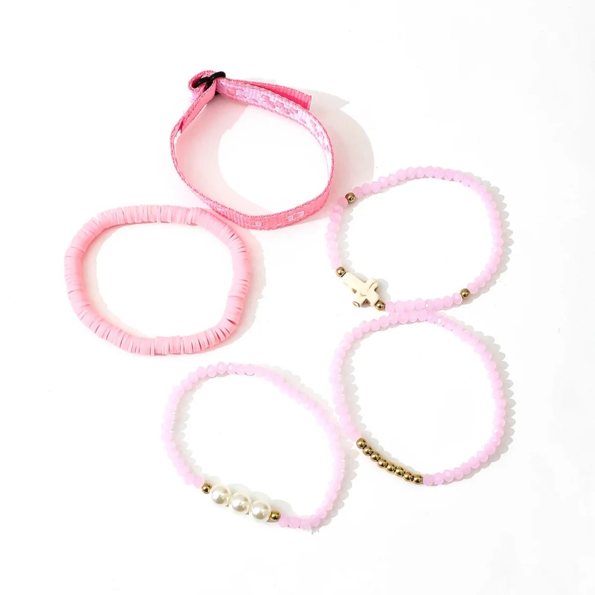Five Kiss Me Later pastel pink bracelets are arranged on a white surface, showcasing unique designs with gold beads, a cross, pearls, woven details, and varied shapes and textures.