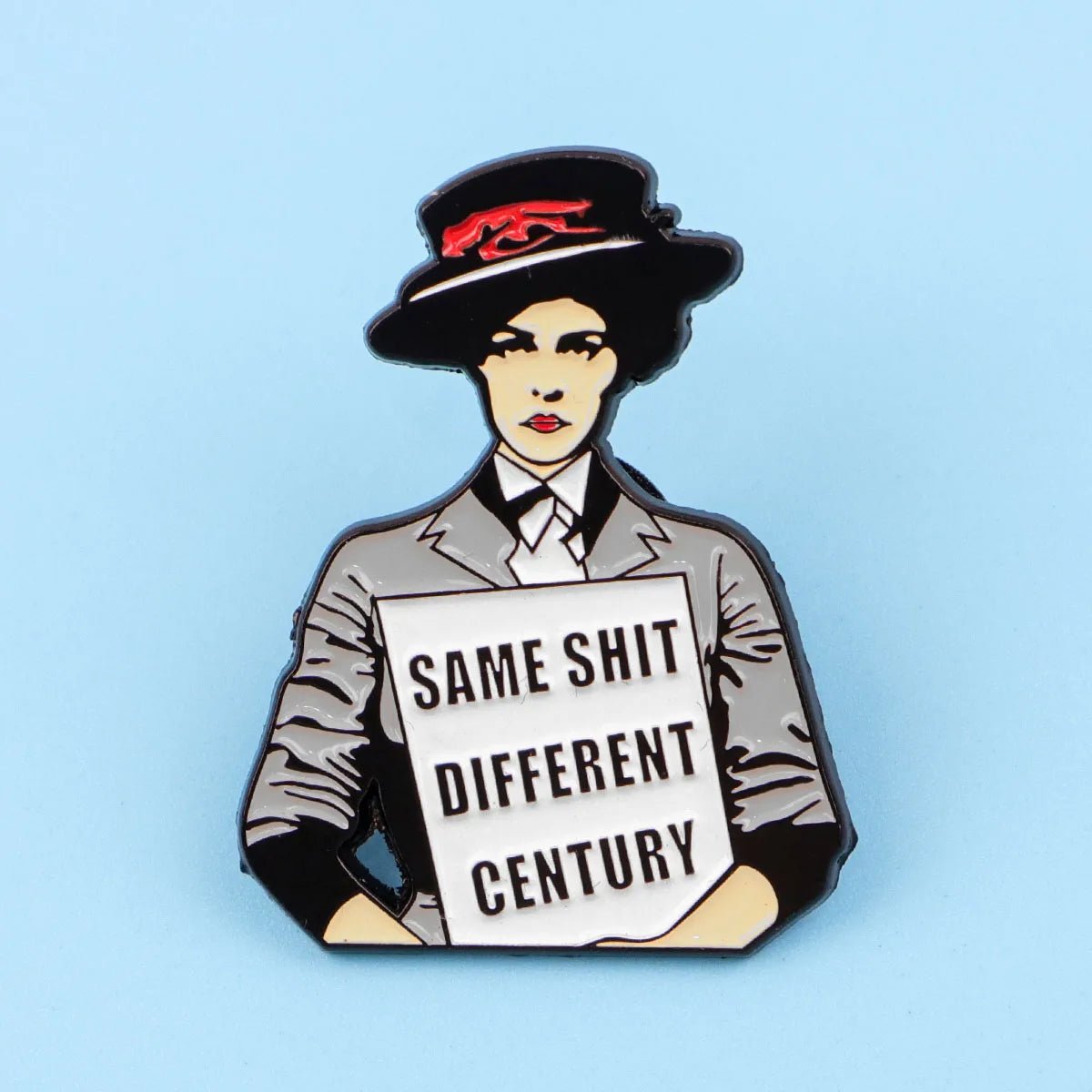 The Kiss Me Later enamel pin features a woman in vintage attire and hat, holding a SAME SHIT DIFFERENT CENTURY sign on a light blue background—a modern twist on the classic suffragette pin.
