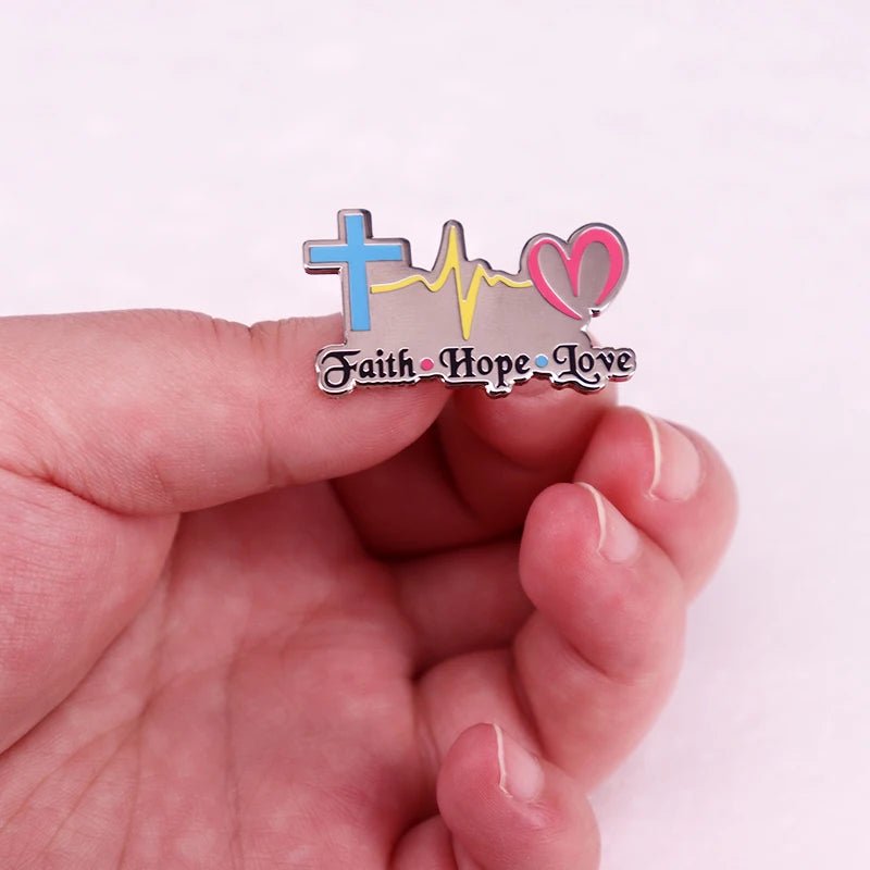 A hand holds a Kiss Me Later badge with a blue cross, yellow heartbeat line, and pink heart above “Faith Hope Love” in decorative script.