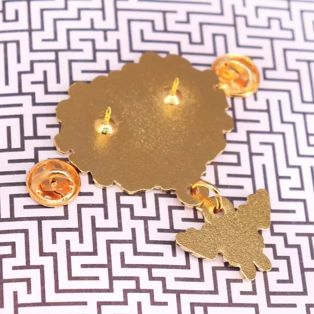 Gold enamel pin features two push-back clasps and a small dangling gold bird, displayed on black and white maze-patterned paper—perfect to pair with your favorite pink and black enamel pins.