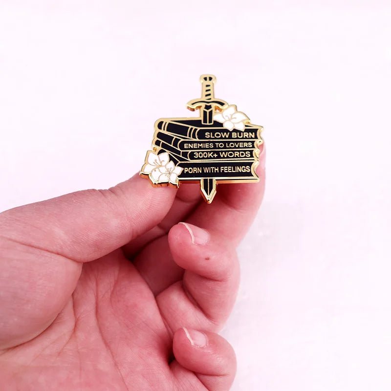 A hand displays an enamel pin showing a stack of books labeled SLOW BURN, ENEMIES TO LOVERS, 300k+ WORDS, and WITH FEELINGS, accented by a sword and white flowers—a must-have for romance trope fans.