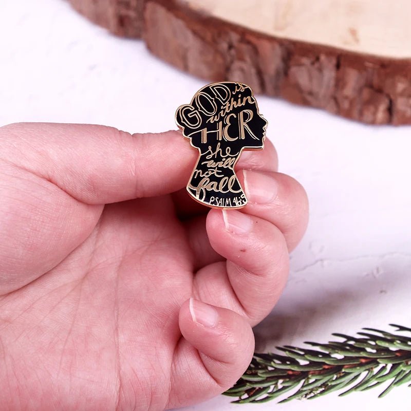 A hand holds a Kiss Me Later God Is Within Her She Will Not Fall enamel pin, featuring a black female silhouette with gold lettering. A pine branch and wood slice appear in the background.