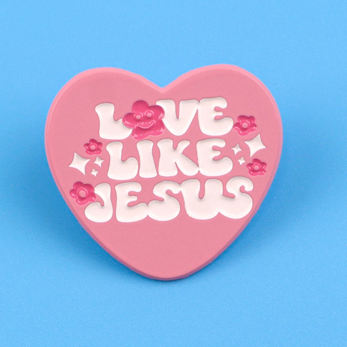 A Kiss Me Later pink love heart pin features LOVE LIKE JESUS in white, with small pink flowers and white sparkles on a blue background.