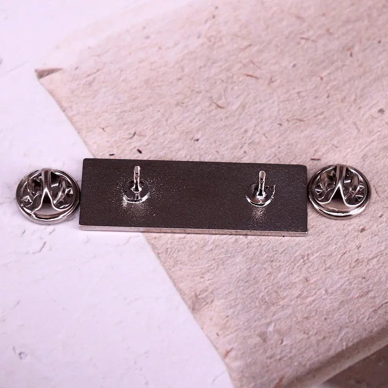 A rectangular black and silver enamel pin with two prongs and circular butterfly clutch backings, displayed on a textured light surface.
