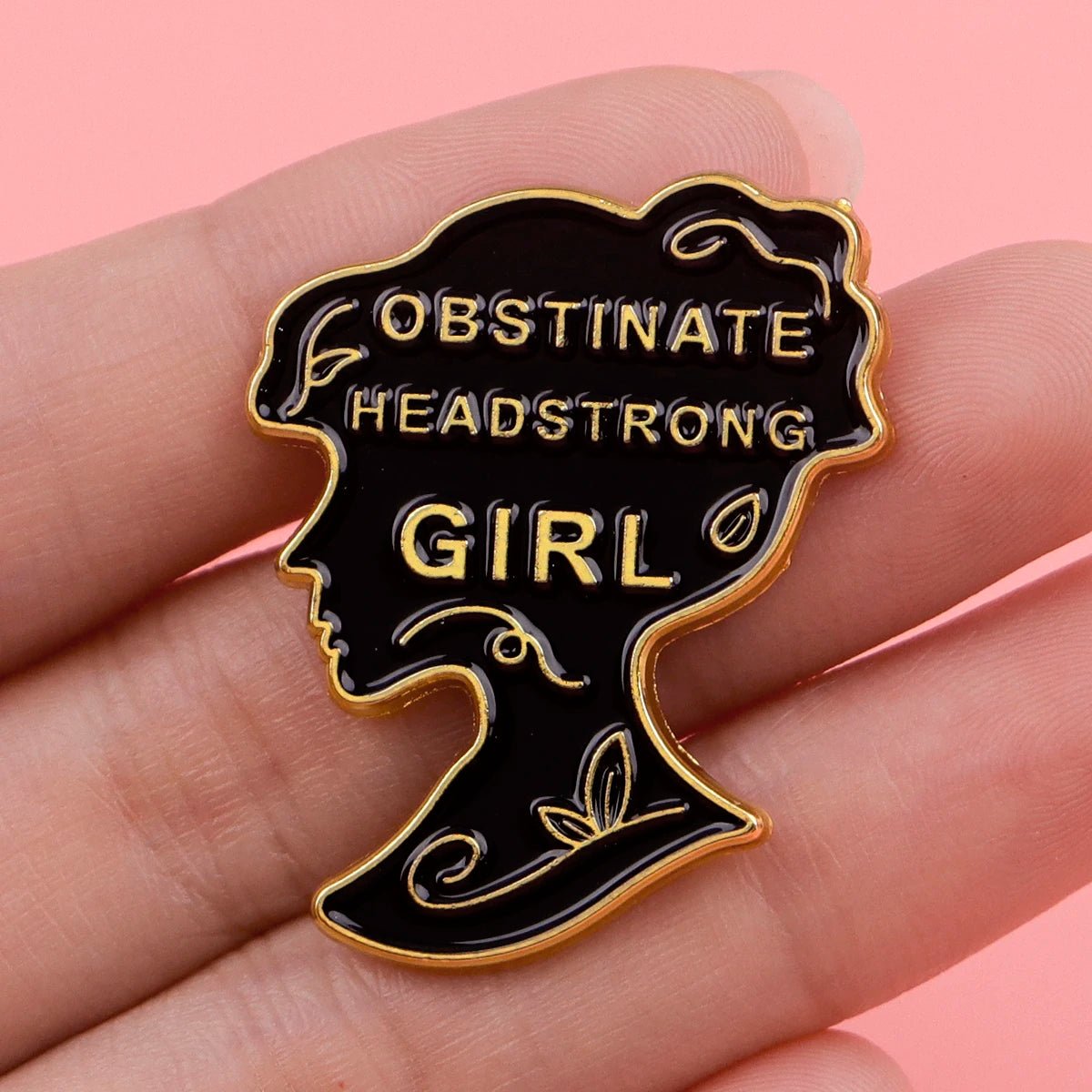 A hand holds a black and gold enamel pin shaped like a girls silhouette with OBSTINATE HEADSTRONG GIRL inside. The pink background highlights the striking pink enamel details on the badge.