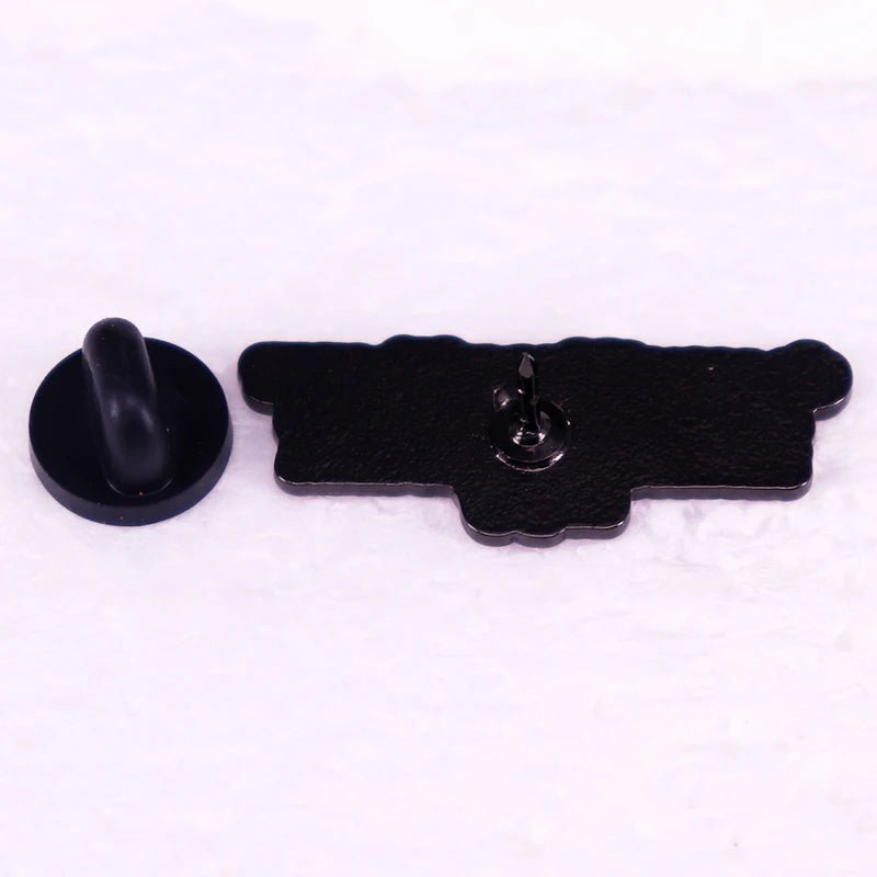 A black enamel cannon-shaped pin from My Cutesy Era lies face down beside a round black rubber pin backing on a white surface.