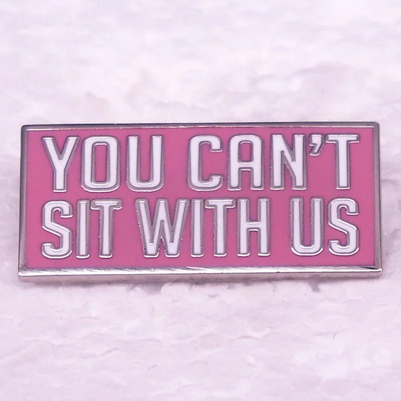 A rectangular pink enamel pin features white and silver text reading YOU CANT SIT WITH US—the perfect accessory for Mean Girls fans, shown on a light textured surface.