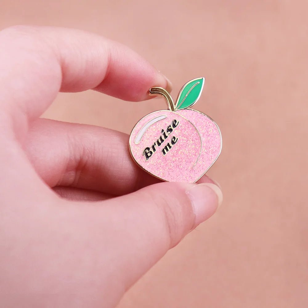 A hand holds a glittery peach enamel pin shaped like a peach with green leaves and the words Bruise Me. The soft peach background complements the playful design.