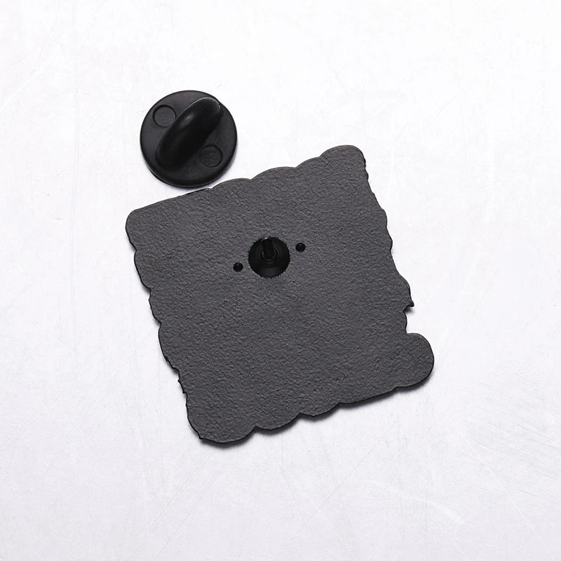 A black, scallop-edged square Jesus pin with a central fastener and two small holes sits on a white surface. The round black pin back is detached and placed above the pin.