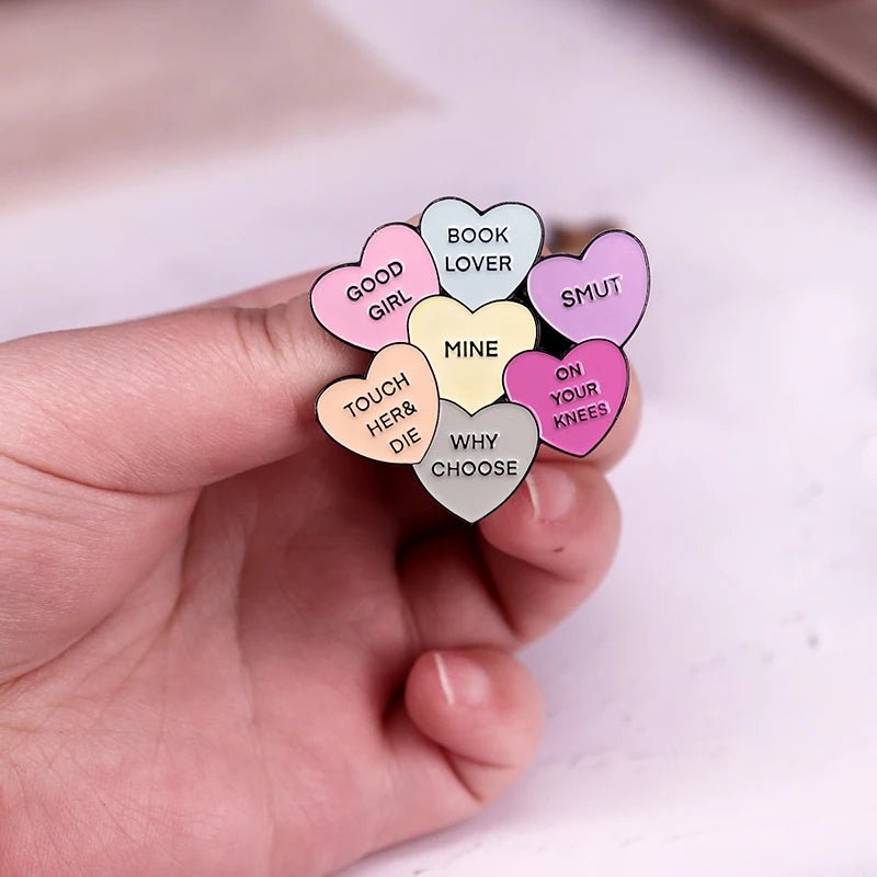 A hand holds an enamel pin shaped like seven pastel conversation hearts with phrases like GOOD GIRL, BOOK LOVER, SMUT, TOUCH HER & DIE, MINE, WHY CHOOSE, and ON YOUR KNEES.