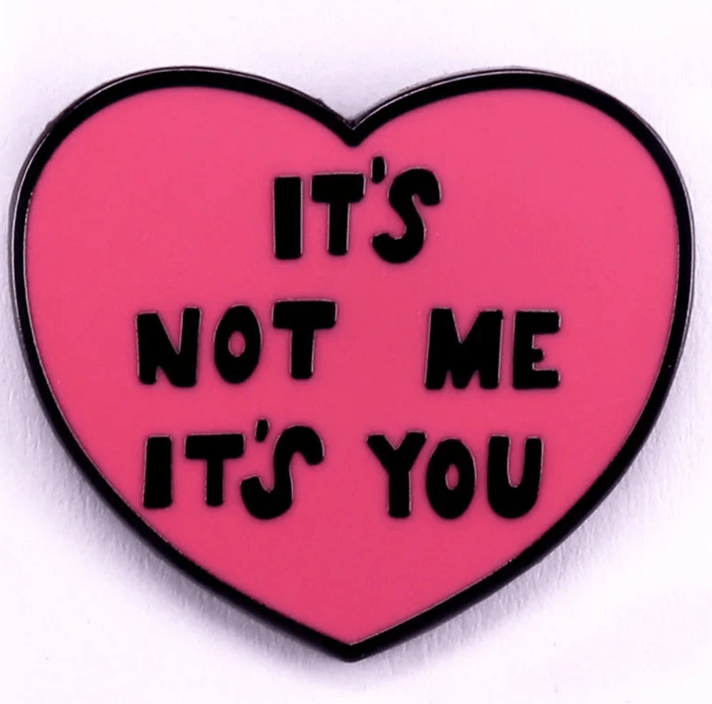Red enamel pin features a pink, heart-shaped design with ITS NOT ME ITS YOU in bold black letters and a striking black outline, making it pop against any background.