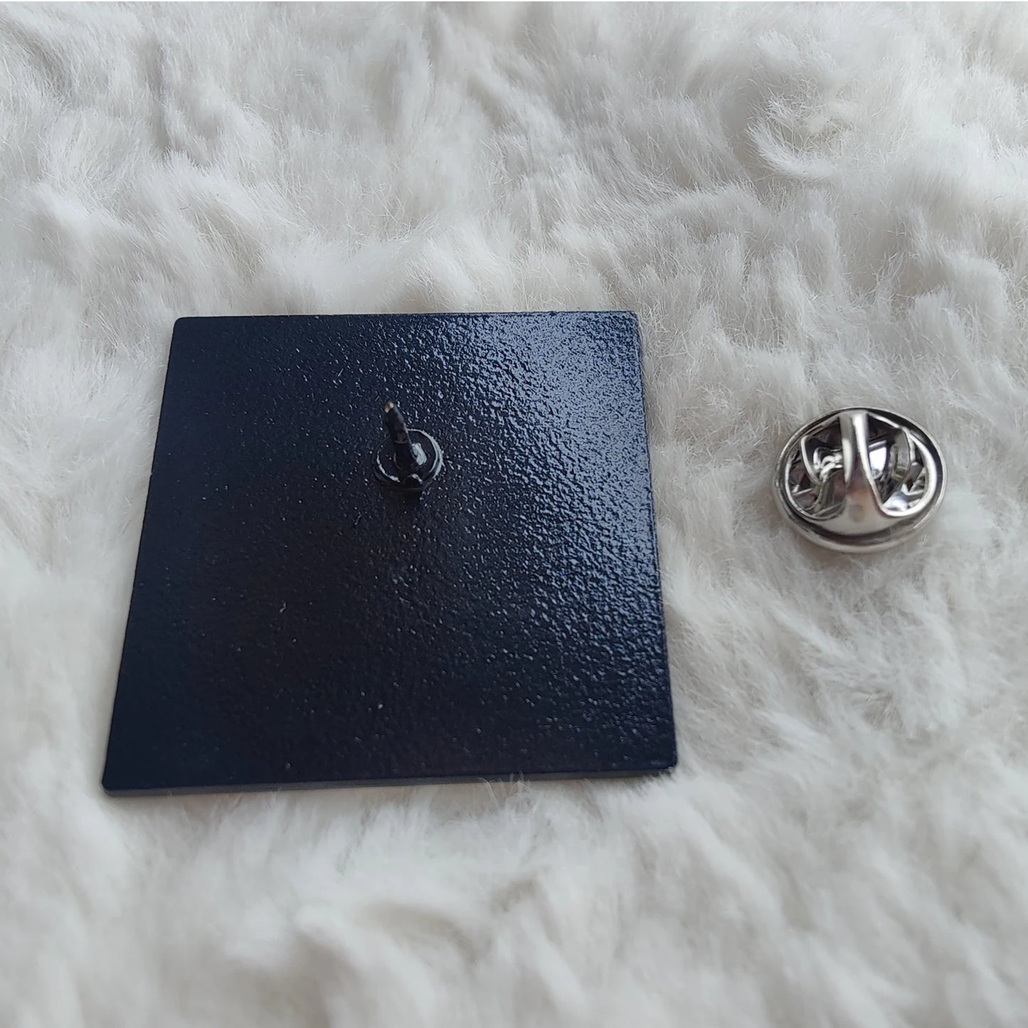 A black, square enamel badge pin by featuring a pointed back and silver butterfly clutch, sits atop a soft white surface—a stylish accessory with Clueless-level charm.