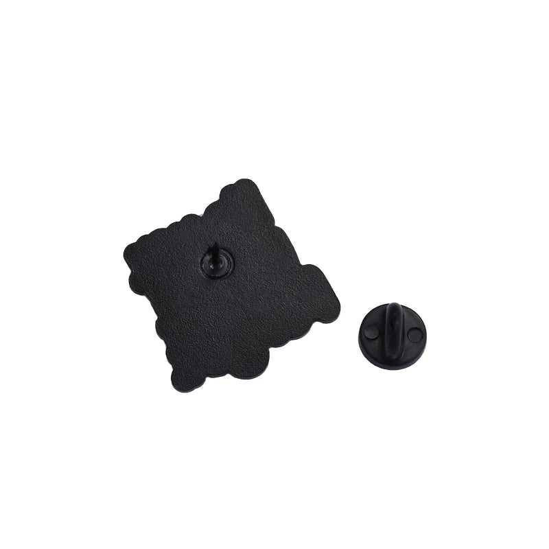 A black enamel pin scalloped square with a textured surface and subtle heart motif, is shown next to its round clasp on a white background.