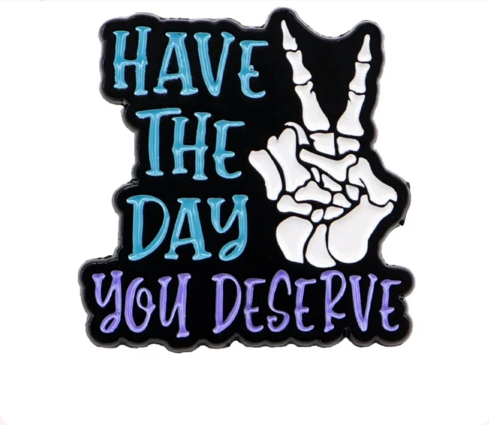 Enamel pin features “Have the day you deserve” in blue and purple with a white skeleton peace sign on a black background, creating a psychedelic vibe.