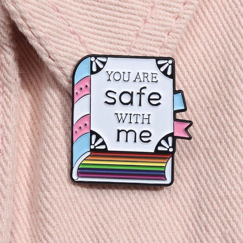 The Kiss Me Later pride enamel pin is shaped like a book with You are safe with me text, features pastel pink, blue, and white trans pride stripes with a rainbow spine, and is attached to pale pink fabric.
