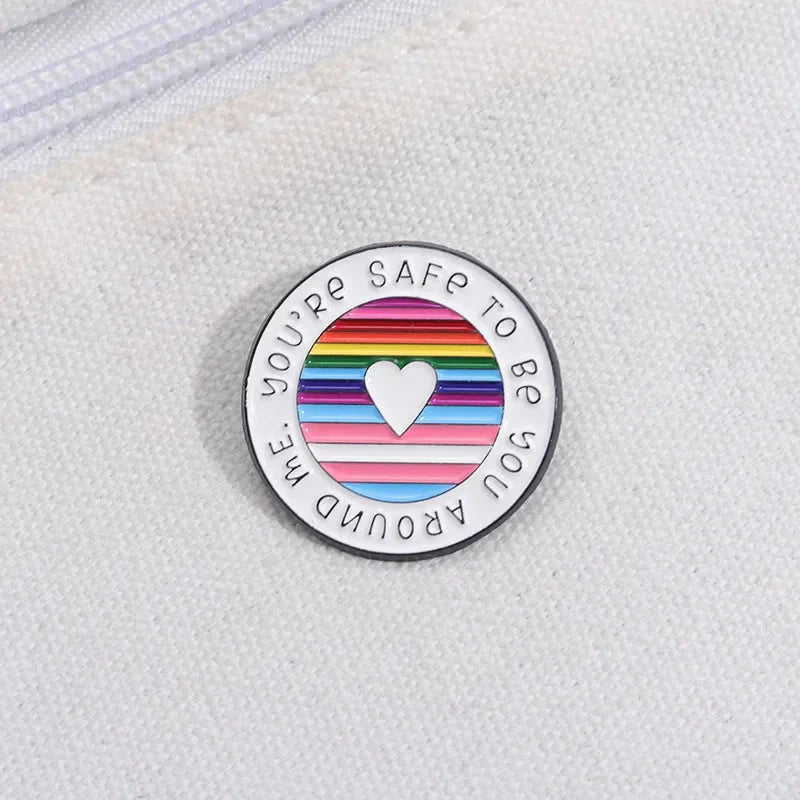 A round enamel pin on white fabric features a white heart over rainbow pride stripes and the message You’re SAFE TO BE YOU AROUND ME circling the edge.