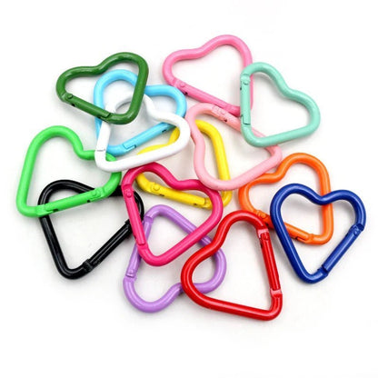 A pile of colorful, heart-shaped rainbow carabiners by My Cutesy Era arranged on a white surface. These vibrant clips, in green, pink, blue, orange, red, black, purple, and white, make the perfect queer accessory.