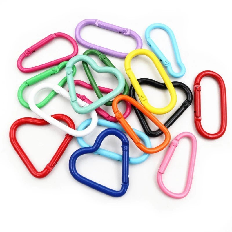 My Cutesy Era offers colorful rainbow carabiners in heart and oval shapes, available in red, blue, yellow, green, pink, purple, white, and black—perfect as a queer accessory or for the wlw community. Scattered on a white surface.