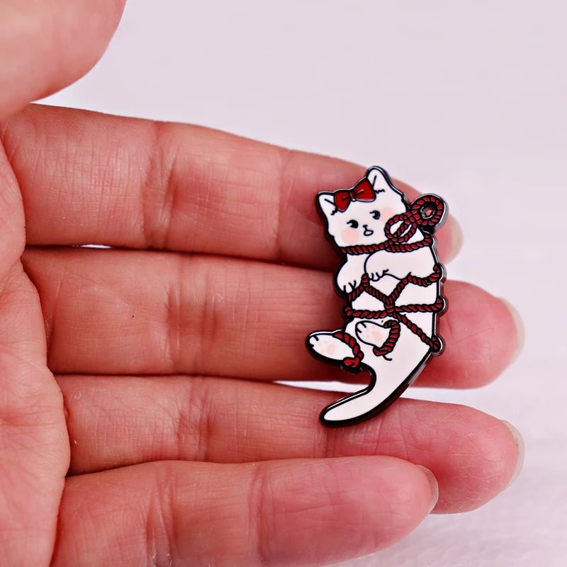 A hand holds an enamel pin featuring a white kitten with a red bow, playfully tangled in red rope.