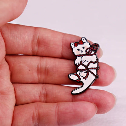A hand holds an enamel pin featuring a white kitten with a red bow, playfully tangled in red rope.