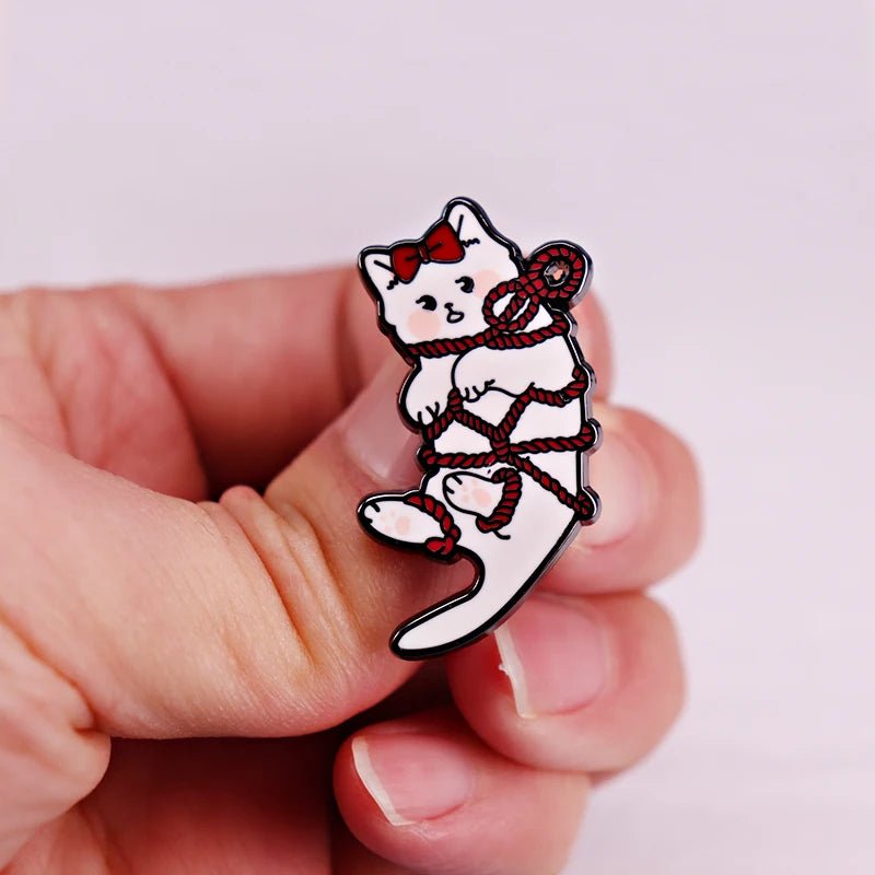 A hand holds an enamel pin showing a white cat with a red bow, playfully wrapped in red rope. The background is light pink.