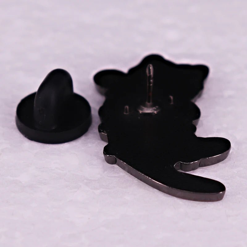 A kitten enamel pin, shaped like a cat, lies face down on a textured white surface with its black rubber backing detached and placed beside it.