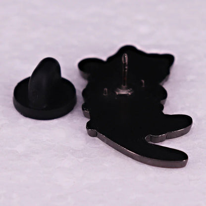 A kitten enamel pin, shaped like a cat, lies face down on a textured white surface with its black rubber backing detached and placed beside it.