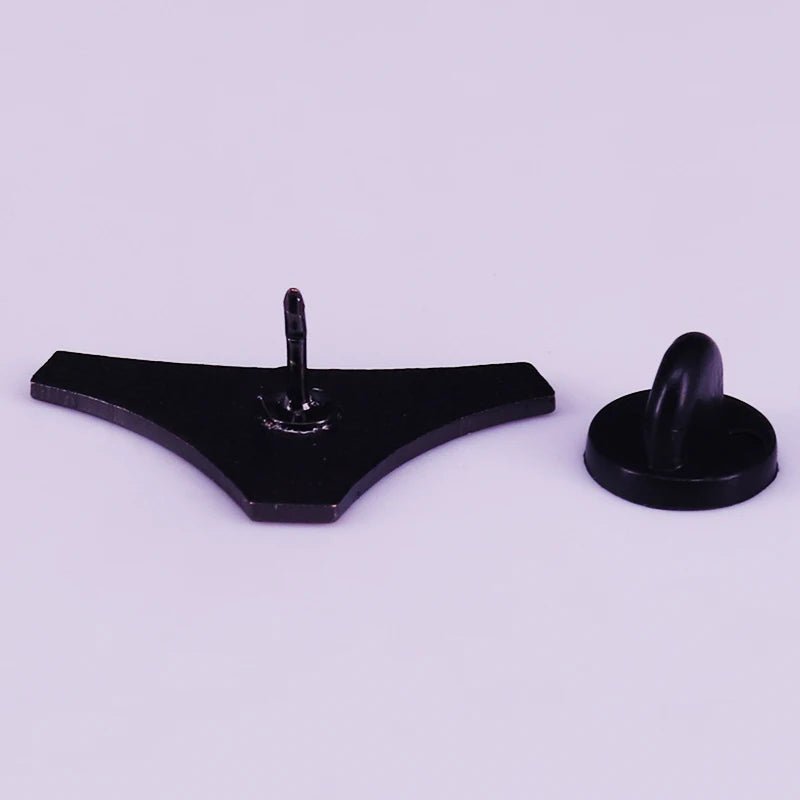 A black, triangular enamel pin with a central peg is positioned to the left of a small, round black knob on a light purple background.