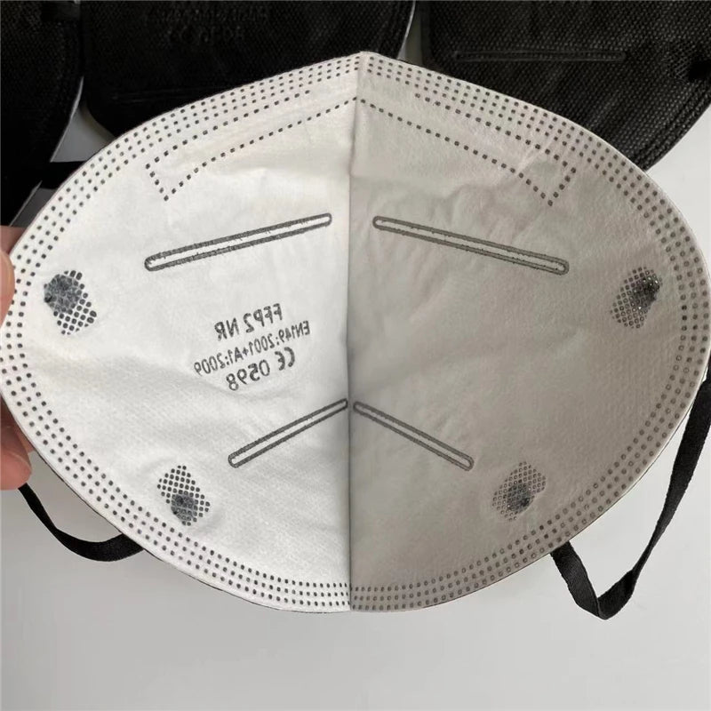 A close-up of a person holding an unfolded white Kiss Me Later FFP2 NR face mask, revealing the inner side with printed certification marks and black elastic ear loops on each side, styled similarly to a KN95 face mask.