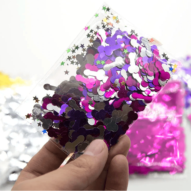 A hand holds a clear Kiss Me Later bag filled with silver and purple metallic penis-shaped confetti mixed with colorful star shapes—fun, cheeky confetti perfect for any adult party. Other vibrant confetti bags are blurred in the background.