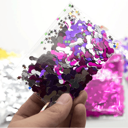 A hand holds a clear Kiss Me Later bag filled with silver and purple metallic penis-shaped confetti mixed with colorful star shapes—fun, cheeky confetti perfect for any adult party. Other vibrant confetti bags are blurred in the background.