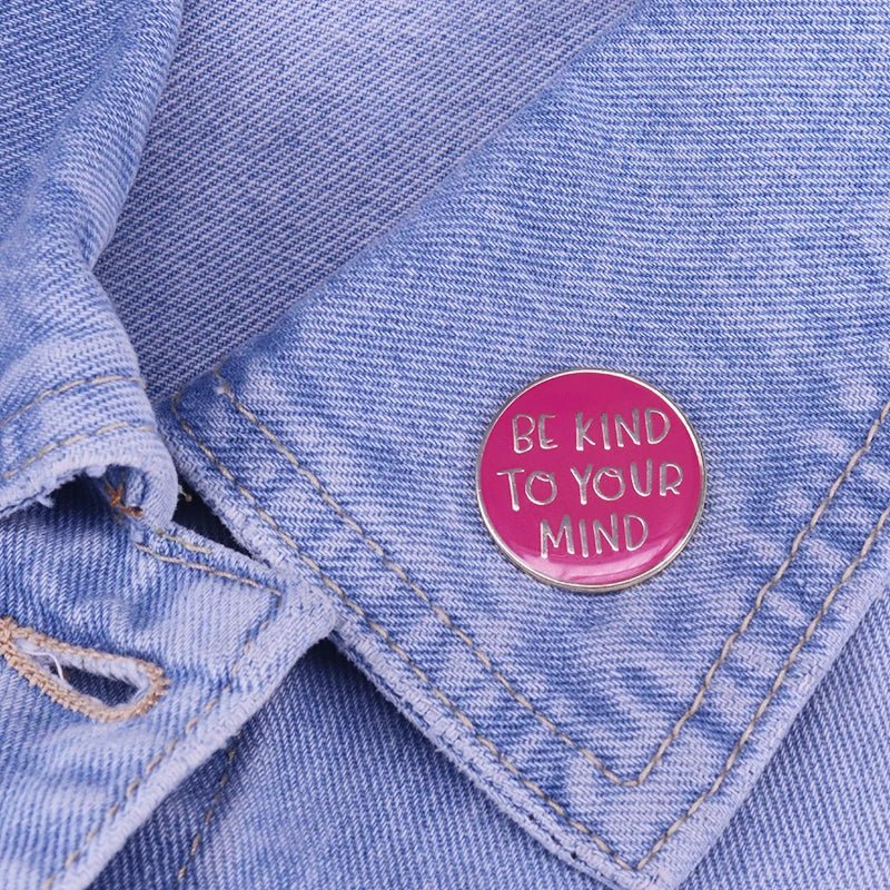 A round pink enamel pin sits on light blue denim, featuring BE KIND TO YOUR MIND in white letters—a meaningful mental health accessory.