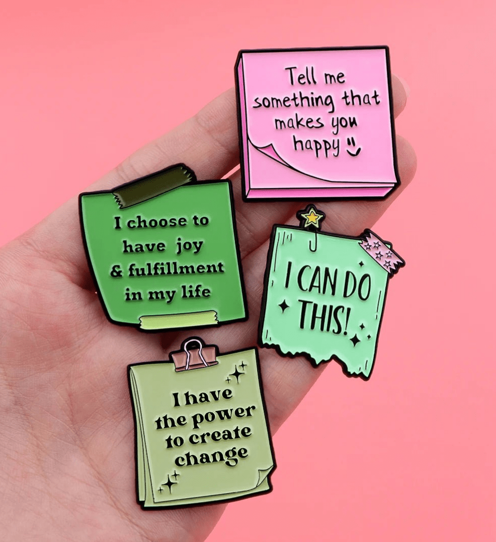 A hand holds four enamel pins shaped like notepaper, each with a motivational message for self-care, including “Tell me something that makes you happy” and “I choose to have joy & fulfillment in my life.”.