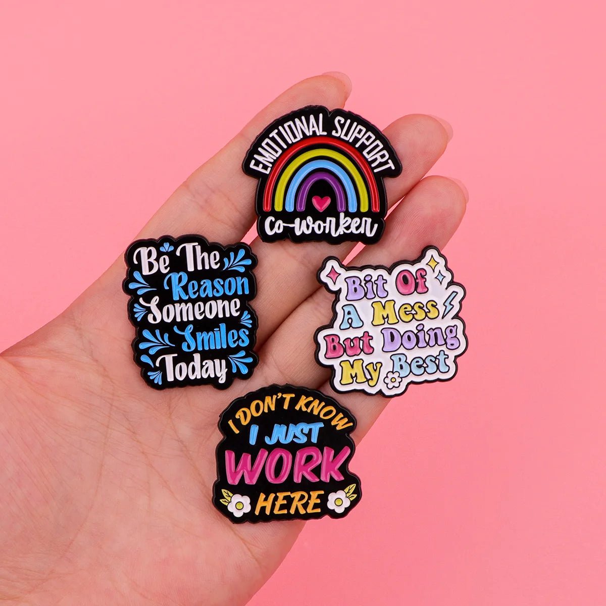 A hand displays four colorful pins on a pink background: Emotional support (rainbow), Be the reason someone smiles today, Bit of a mess but doing my best, and I don’t know I just work here—perfect coworker gifts.