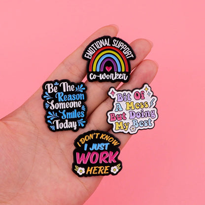 A hand displays four colorful pins on a pink background: Emotional support (rainbow), Be the reason someone smiles today, Bit of a mess but doing my best, and I don’t know I just work here—perfect coworker gifts.