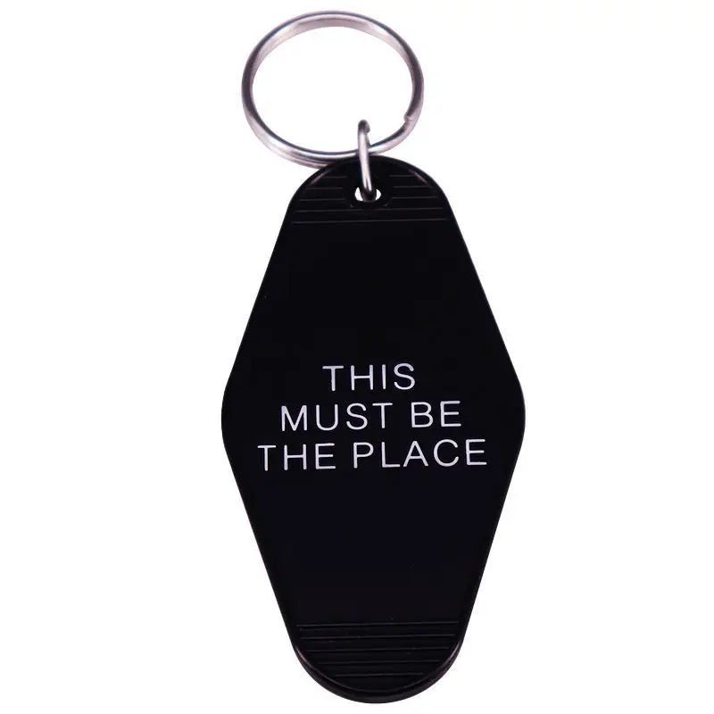 A black keychain by Kiss Me Later with a silver ring, inspired by retro hotel key tags, features THIS MUST BE THE PLACE in bold white capitals.