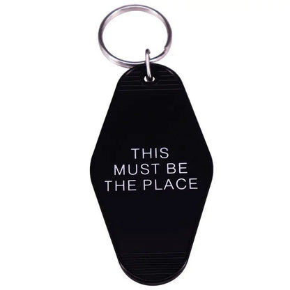 A black keychain by Kiss Me Later with a silver ring, inspired by retro hotel key tags, features THIS MUST BE THE PLACE in bold white capitals.