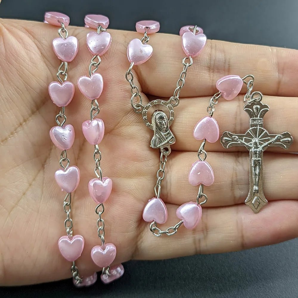 A hand holds a My Cutesy Era Rosary necklace featuring pink heart-shaped beads, a Virgin Mary medal, and a silver crucifix against a dark background.