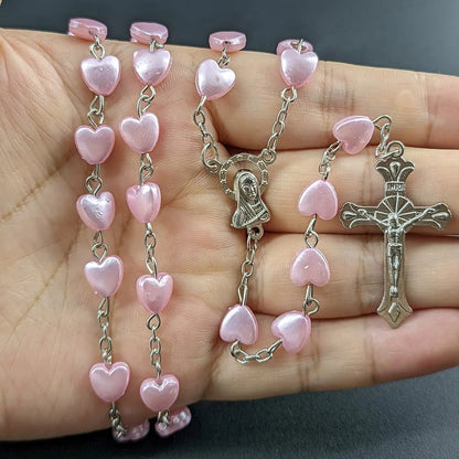 A hand holds a My Cutesy Era Rosary necklace featuring pink heart-shaped beads, a Virgin Mary medal, and a silver crucifix against a dark background.