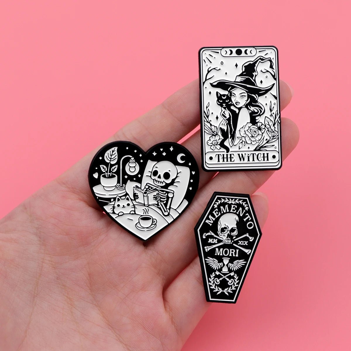 A hand displays three Kiss Me Later enamel pins: a heart-shaped skeleton hand having tea, a rectangular THE WITCH pin with a cat, and a coffin-shaped MEMENTO MORI skull pin—all in spooky black and white.