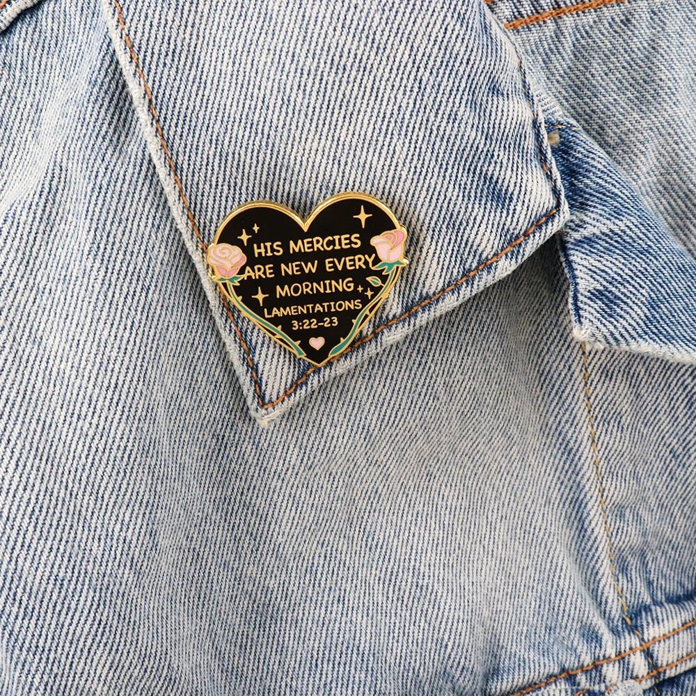 A heart-shaped enamel pin on a denim jacket reads, “His Mercies Are New every morning. Lamentations 3:22-23,” and features two pink roses with gold accents.