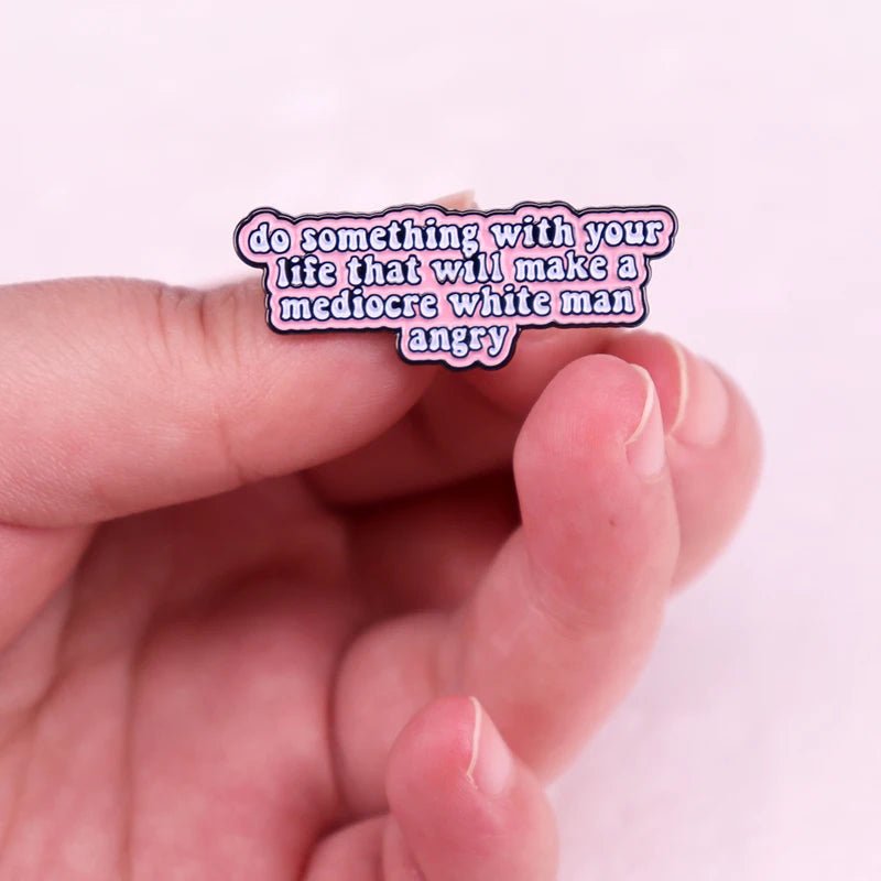 A hand holds a My Cutesy Era feminist enamel pin with the text: do something with your life that will make a mediocre white man angry, shown on a pink background.