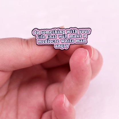 A hand holds a My Cutesy Era feminist enamel pin with the text: do something with your life that will make a mediocre white man angry, shown on a pink background.