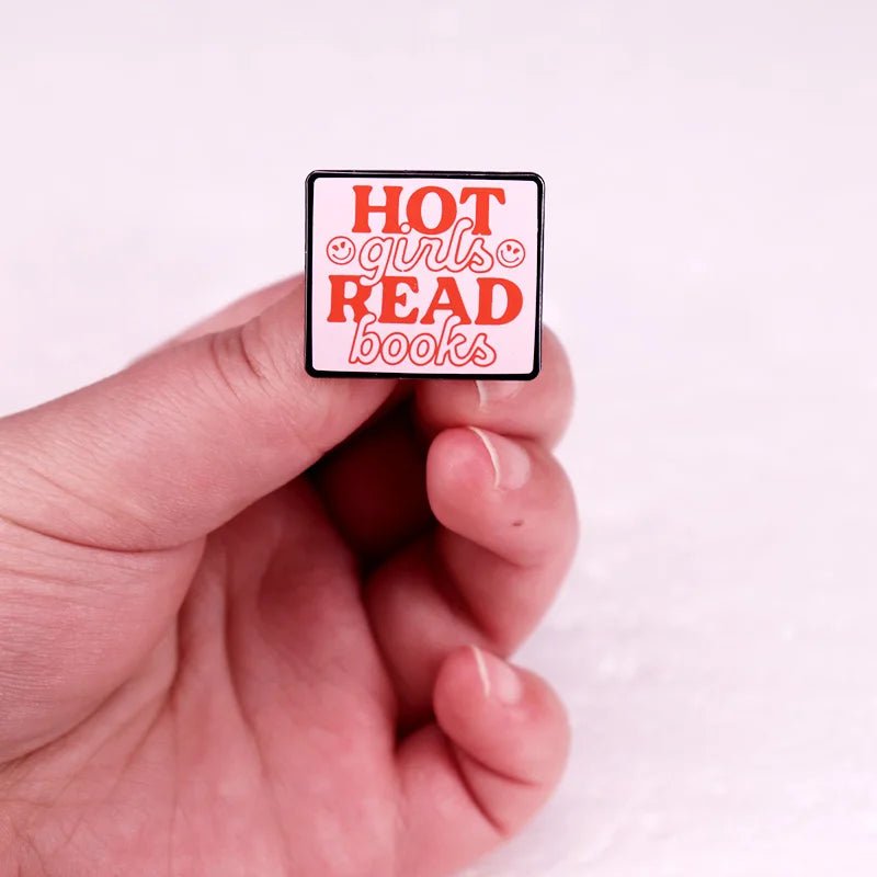A hand holds a rectangular Kiss Me Later Hot Girls Read Books enamel pin with bold red and orange letters on a light background—an ideal book lover pin for any passionate reader.