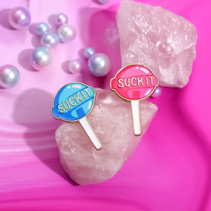 Two Suck It pins, shaped like lollipops in blue and pink enamel, rest on a pink crystal amid pearl-like beads—adding playful charm to any collection.