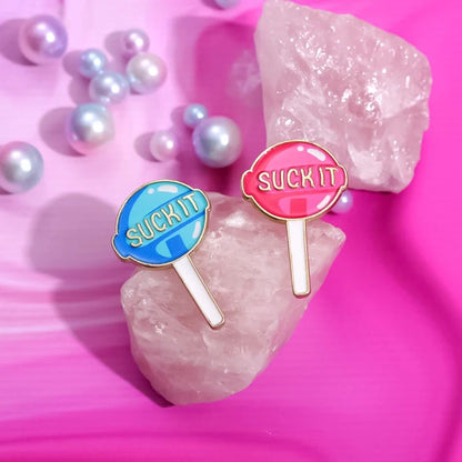 Two Suck It pins, shaped like lollipops in blue and pink enamel, rest on a pink crystal amid pearl-like beads—adding playful charm to any collection.